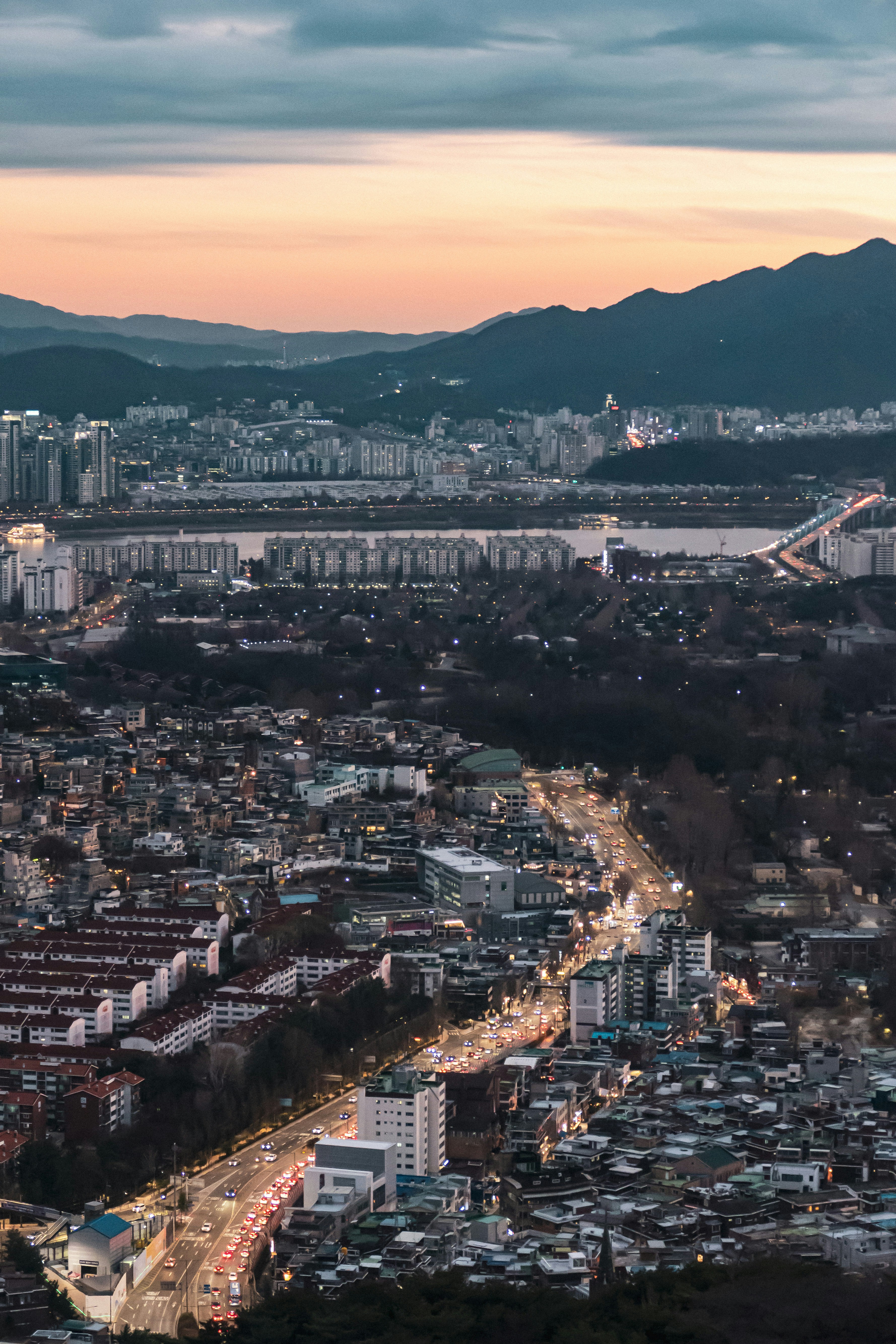 Cityscape photograph at dusk showing a glowing highway winding through dense housing beside a river. Distant mountains silhouette the skyline.