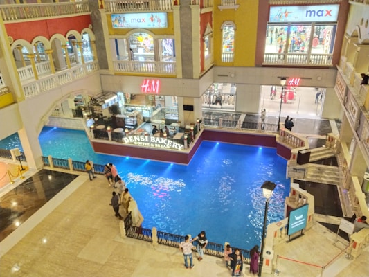 A multi-story shopping mall with indoor balconies overlooking a spacious area featuring a central water body, bordered by a bakery (Densi Bakery Coffee & Bread) and several retail stores, including H&M and Max. Shoppers are seen walking around and standing near the water, and the area is well-lit with a blend of natural and artificial lights.