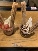 Two cups of soft serve ice cream are garnished with long churros. One cup is topped with chocolate sauce, and the other with strawberry pieces.