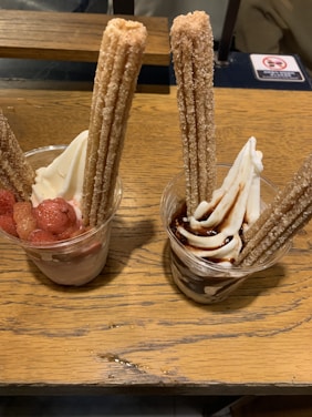 Two cups of soft serve ice cream are garnished with long churros. One cup is topped with chocolate sauce, and the other with strawberry pieces.