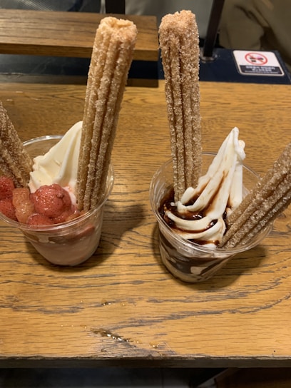 Two cups of soft serve ice cream are garnished with long churros. One cup is topped with chocolate sauce, and the other with strawberry pieces.