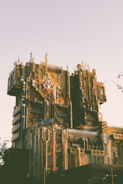 A large, intricate building with a futuristic design featuring angular lines, metallic elements, and various textures. The structure is adorned with a mix of geometric patterns and mechanical details, creating a complex facade. The lighting suggests either a sunset or sunrise, casting a warm glow over the building.