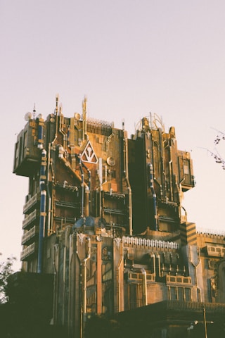A large, intricate building with a futuristic design featuring angular lines, metallic elements, and various textures. The structure is adorned with a mix of geometric patterns and mechanical details, creating a complex facade. The lighting suggests either a sunset or sunrise, casting a warm glow over the building.