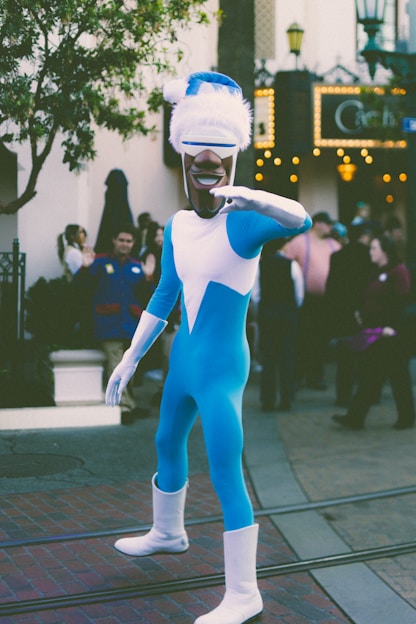 A person dressed in a bright blue and white superhero costume poses on a street. The costume includes a full-body suit with a headpiece and sunglasses. There are people in the background on a brick-paved street, and a tree is visible to the left. Decorative lights adorn a building in the background.