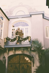 An elegant building facade adorned with intricate architectural details and a decorative balcony displaying a festive garland with gold ribbons. A statue is prominently positioned on the balcony, while palm fronds add a touch of greenery.