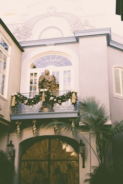 An elegant building facade adorned with intricate architectural details and a decorative balcony displaying a festive garland with gold ribbons. A statue is prominently positioned on the balcony, while palm fronds add a touch of greenery.