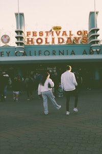 Two people walk towards a building entrance adorned with a sign that reads 'Festival of Holidays' with 'California Adventure' visible below. There are other visitors in the vicinity, and the lighting suggests it might be late afternoon or early evening.
