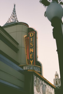 A building with an art deco style features a vertical neon sign that reads 'Disney Theater'. It has intricate architectural details, including a decorative spire atop the building. A palm tree and what appears to be an ornate street lamp are partially visible.