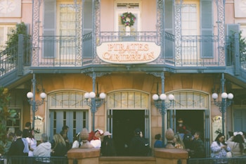A group of people stand in line outside an entrance decorated with a sign reading 'Pirates of the Caribbean.' The building features intricate wrought iron railings and a festive wreath above the sign. Architectural details include beige walls and blue shutters.