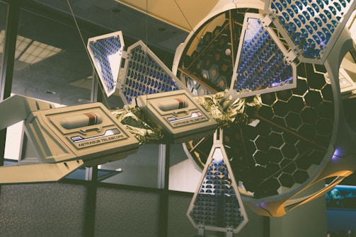 A sophisticated, futuristic telescope installation with hexagonal patterns and blue and gold detailing is displayed indoors. The structure features solar panel-like components and intricate mechanical elements, suggesting advanced technology and scientific innovation.