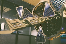 A sophisticated, futuristic telescope installation with hexagonal patterns and blue and gold detailing is displayed indoors. The structure features solar panel-like components and intricate mechanical elements, suggesting advanced technology and scientific innovation.