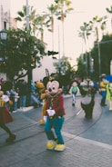A lively parade features popular cartoon characters walking along a street flanked by spectators. Tall palm trees and urban architecture provide a backdrop to the colorful scene.