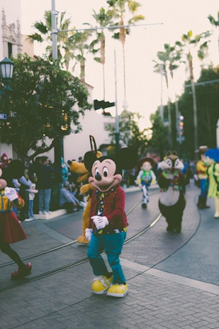A lively parade features popular cartoon characters walking along a street flanked by spectators. Tall palm trees and urban architecture provide a backdrop to the colorful scene.