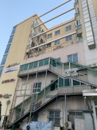 A multi-story building with a beige and glass facade features several balconies and external staircases leading to different floors. Signage with Korean characters is visible on the walls, and there are stacked water bottles and various equipment at ground level. The sky is clear and blue, suggesting daylight.