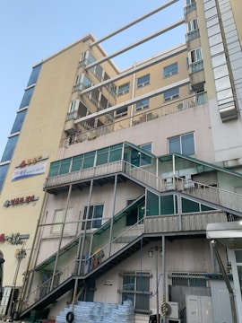 A multi-story building with a beige and glass facade features several balconies and external staircases leading to different floors. Signage with Korean characters is visible on the walls, and there are stacked water bottles and various equipment at ground level. The sky is clear and blue, suggesting daylight.