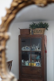 An antique wooden cabinet with glass doors displays various household items, including framed photographs, ceramics, and glassware. On top of the cabinet, a wicker basket with green foliage rests alongside another basket and a brass candlestick holder. The scene is reflected in a decorative ornate mirror.