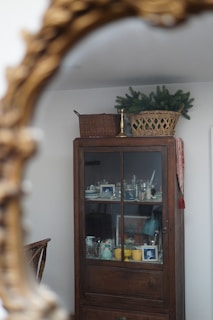 An antique wooden cabinet with glass doors displays various household items, including framed photographs, ceramics, and glassware. On top of the cabinet, a wicker basket with green foliage rests alongside another basket and a brass candlestick holder. The scene is reflected in a decorative ornate mirror.
