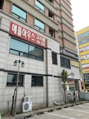 A multi-story building with a gray and pink facade is visible with signs in Korean script. The ground level features shop signs and air conditioning units attached to the exterior. There are parking spaces and street lamps in the foreground.