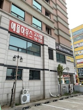 A multi-story building with a gray and pink facade is visible with signs in Korean script. The ground level features shop signs and air conditioning units attached to the exterior. There are parking spaces and street lamps in the foreground.