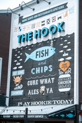 A mural on the side of a building advertising a seafood restaurant named 'The Hook.' The design includes nautical elements such as flags and a large fish, with various humorous and enticing phrases promoting items like fish and chips, crab, shrimp chowder, and grilled chicken sandwiches. The mural uses blues, whites, and blacks to create a sea-related theme.