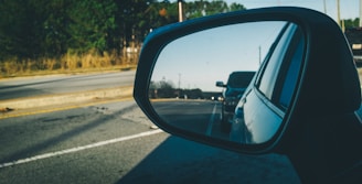 A side-view mirror of a car showing the reflection of another car behind it, driving on a road bordered by trees and vegetation. The mirror has an indicator light or symbol.