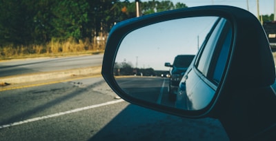 A side-view mirror of a car showing the reflection of another car behind it, driving on a road bordered by trees and vegetation. The mirror has an indicator light or symbol.