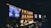 A dimly lit bar with a modern design features bottles of liquor displayed on illuminated shelves. A large television screen is mounted to the left, showing a person speaking with subtitles. The bar counter is adorned with candles and various glassware. Two other screens in the background display different logos or images, contributing to the ambient lighting.
