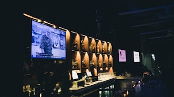 A dimly lit bar with a modern design features bottles of liquor displayed on illuminated shelves. A large television screen is mounted to the left, showing a person speaking with subtitles. The bar counter is adorned with candles and various glassware. Two other screens in the background display different logos or images, contributing to the ambient lighting.