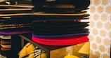 A stack of various colored hats, including black, red, and blue, is neatly arranged on a display shelf. There is a patterned fabric visible in the lower section, adding to the organized appearance. The background features a geometric, circular pattern in white against a beige backdrop.