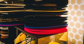 A stack of various colored hats, including black, red, and blue, is neatly arranged on a display shelf. There is a patterned fabric visible in the lower section, adding to the organized appearance. The background features a geometric, circular pattern in white against a beige backdrop.