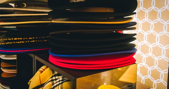 A stack of various colored hats, including black, red, and blue, is neatly arranged on a display shelf. There is a patterned fabric visible in the lower section, adding to the organized appearance. The background features a geometric, circular pattern in white against a beige backdrop.