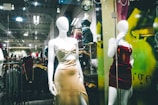 Mannequins in a store display window feature elegant dresses. The setting appears to be a modern clothing store with a sleek design, visible through reflections on the glass. The dresses are stylish and party-ready, with satin textures and bold colors. Background details suggest a bustling shopping area with clothing racks and ambient lighting.