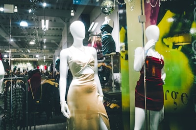Mannequins in a store display window feature elegant dresses. The setting appears to be a modern clothing store with a sleek design, visible through reflections on the glass. The dresses are stylish and party-ready, with satin textures and bold colors. Background details suggest a bustling shopping area with clothing racks and ambient lighting.