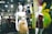 Mannequins in a store display window feature elegant dresses. The setting appears to be a modern clothing store with a sleek design, visible through reflections on the glass. The dresses are stylish and party-ready, with satin textures and bold colors. Background details suggest a bustling shopping area with clothing racks and ambient lighting.