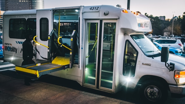 A transit shuttle bus with an extended wheelchair ramp is parked on a street. The bus is marked with 'MARTA Mobility' and is designed for accessibility. The surroundings include a parking lot with numerous cars under a dusky sky.