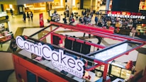 A bustling food court inside a shopping mall featuring a CamiCakes Cupcakes kiosk with its prominent signage in the foreground. In the background, several people are walking and sitting around tables, with a Starbucks Coffee shop visible. The interior is well-lit with a warm color palette, and various retail stores and kiosks are scattered throughout the space.