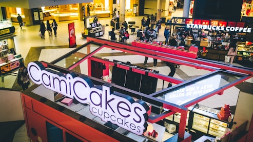 A bustling food court inside a shopping mall featuring a CamiCakes Cupcakes kiosk with its prominent signage in the foreground. In the background, several people are walking and sitting around tables, with a Starbucks Coffee shop visible. The interior is well-lit with a warm color palette, and various retail stores and kiosks are scattered throughout the space.