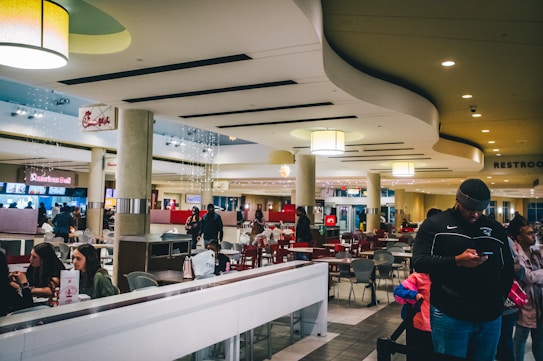 A spacious and modern food court with various seating areas and people interacting. Overhead lighting and clean design elements create a bright and welcoming atmosphere. There are several food stalls, including recognizable chains, and a few people are seen waiting or on their phones.