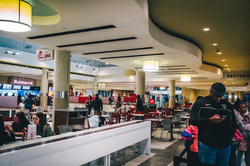 A spacious and modern food court with various seating areas and people interacting. Overhead lighting and clean design elements create a bright and welcoming atmosphere. There are several food stalls, including recognizable chains, and a few people are seen waiting or on their phones.