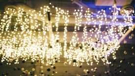 A ceiling installation featuring numerous small spherical lights, creating a sparkling and twinkling effect. The lights are scattered irregularly, casting a warm glow against the backdrop. The setting appears to be an indoor space, possibly a modern venue or art installation, with additional architectural details visible above the light arrangement.