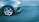 The image captures the front left part of a silver car parked on an asphalt road. The car's wheel and headlight are clearly visible, and the road's texture and a faint white line in the background are also noticeable.