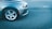The image captures the front left part of a silver car parked on an asphalt road. The car's wheel and headlight are clearly visible, and the road's texture and a faint white line in the background are also noticeable.