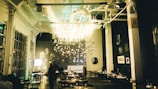 A warmly lit restaurant interior features modern decor with hanging light installations and a Christmas tree on the side. The dining area has neatly arranged tables and chairs, and the walls display various artworks. The ambiance is cozy and inviting.