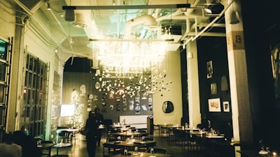 A warmly lit restaurant interior features modern decor with hanging light installations and a Christmas tree on the side. The dining area has neatly arranged tables and chairs, and the walls display various artworks. The ambiance is cozy and inviting.
