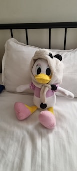 A plush toy resembling a duck character, wearing a panda-themed hoodie, sits upright on a neatly made bed with white linens. Its body is predominantly white with accents of pink on the feet and chest, and a bright yellow beak.