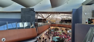 A large, modern indoor space resembling an airport terminal or shopping mall. The architecture features a prominent use of glass and metal, with a curving wooden structure. The lower level is bustling with people, and there are informational signs and various seating areas surrounded by plants.