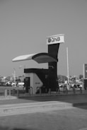 A modern ATM kiosk with the QNB logo is situated in an open area, possibly a parking lot or a walkway. The structure has a sleek design with a prominent canopy and protective barriers around it. In the background, there are multiple parked vehicles and some buildings or structures.