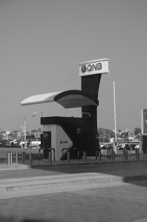 A modern ATM kiosk with the QNB logo is situated in an open area, possibly a parking lot or a walkway. The structure has a sleek design with a prominent canopy and protective barriers around it. In the background, there are multiple parked vehicles and some buildings or structures.