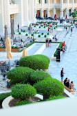 An outdoor patio area with numerous people sitting, walking, and socializing. There are multiple levels of seating with tables and chairs, bordered by neatly trimmed hedges and shrubs. The background features a large building with columns and a retail area. Several umbrellas are open, providing shade for the customers.