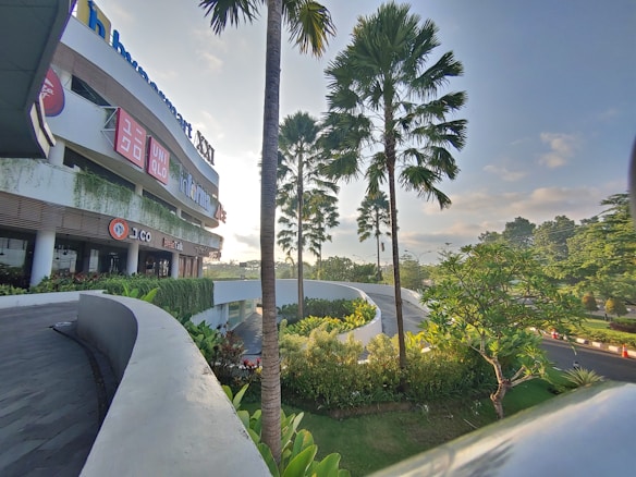 A modern commercial building with multiple store signs including Uniqlo, surrounded by lush greenery and tall palm trees. The sun is setting in the background, casting a warm glow over the curved driveway that leads to the building.